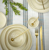 Table setting with beige plates, gold cutlery, and greenery on a striped tablecloth.