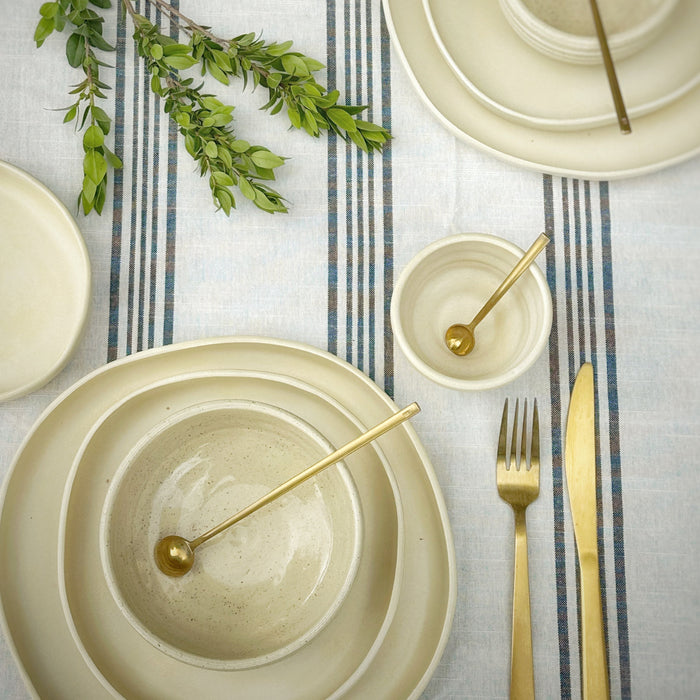 Table setting with beige plates, gold cutlery, and greenery on a striped tablecloth.